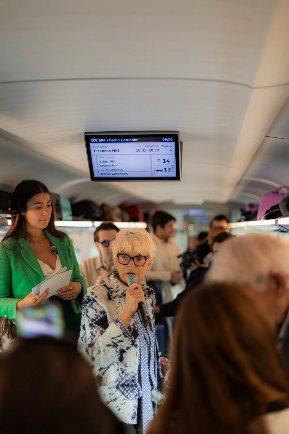 Eine Frau spricht ins Mikrofon, daneben steht eine Moderatorin mit Karten; darüber ein Fahrgastmonitor mit der Zuglaufanzeige nach Berlin.