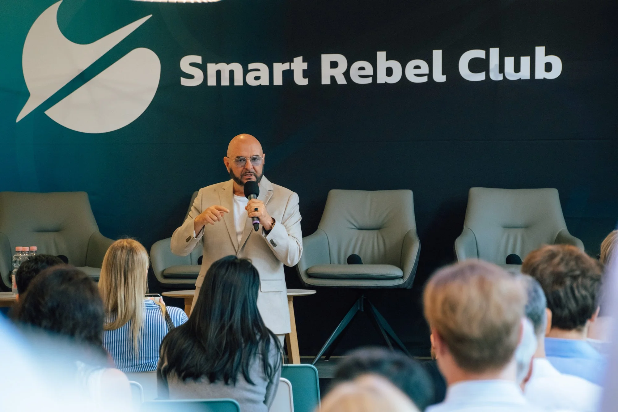 Ein Redner mit Mikrofon steht vor dem Smart-Rebel-Club-Logo, davor leere Sessel und Publikum.