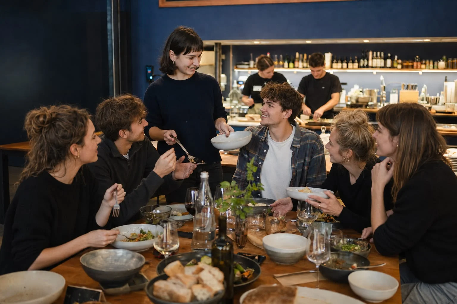 Junge Menschen sitzen beim gemeinsamen Essen um einen langen Holztisch und reichen einander Schüsseln, im Hintergrund eine offene Küche