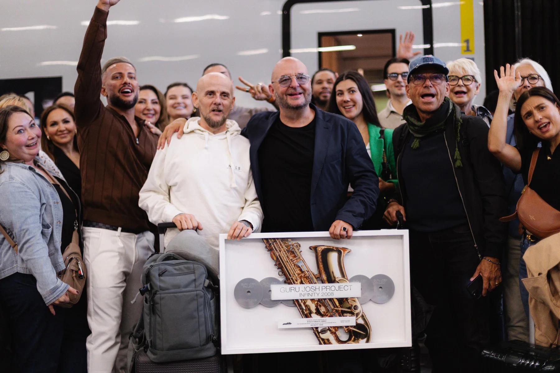 Eine jubelnde Reisegruppe steht auf dem Bahnsteig neben dem ICE und hält eine gerahmte Auszeichnung mit goldenem Saxofon in die Kamera; auf dem Schild stehen „Guru Josh Project“ und „Infinity 2008“.
