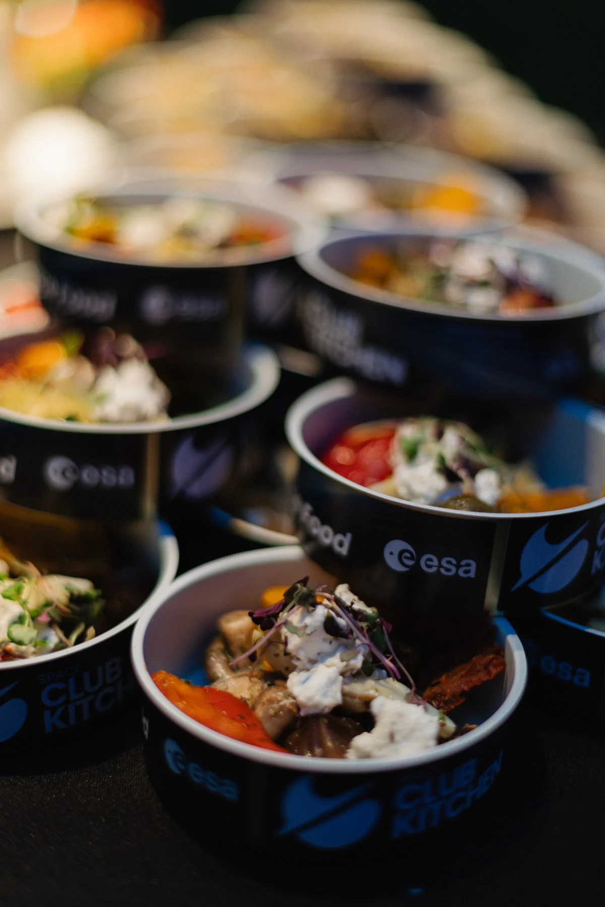 Mehrere Becher mit den Aufschriften Space Club Kitchen und esa, gefüllt mit Salat, Tomaten und Frischkäse.