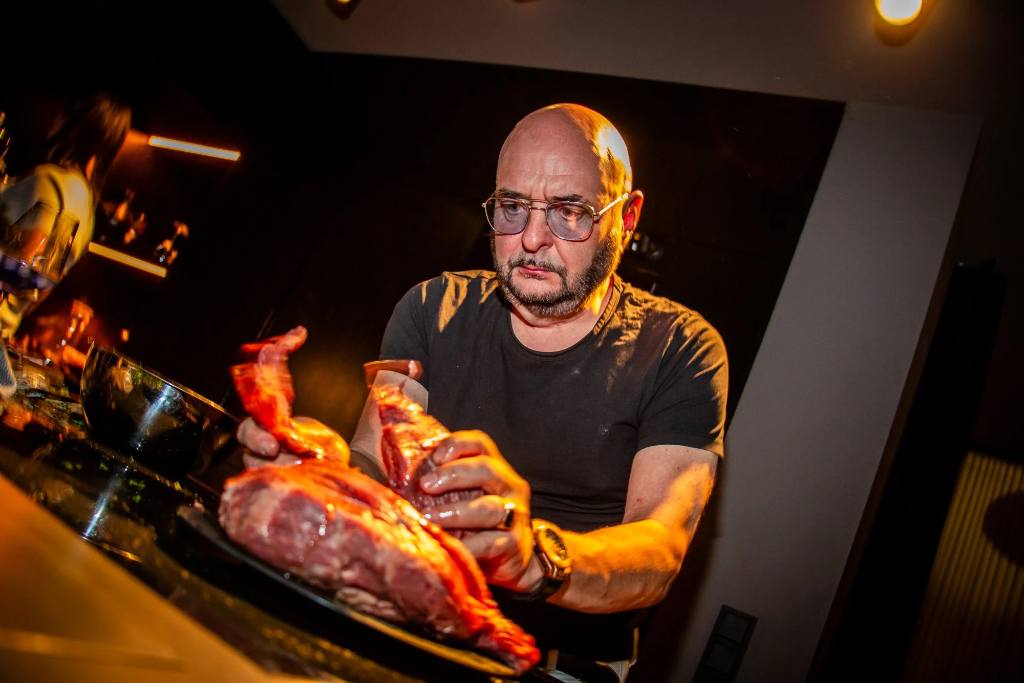 Ein Mann bearbeitet unter warmem Licht ein großes Stück rohes Fleisch am Herd.