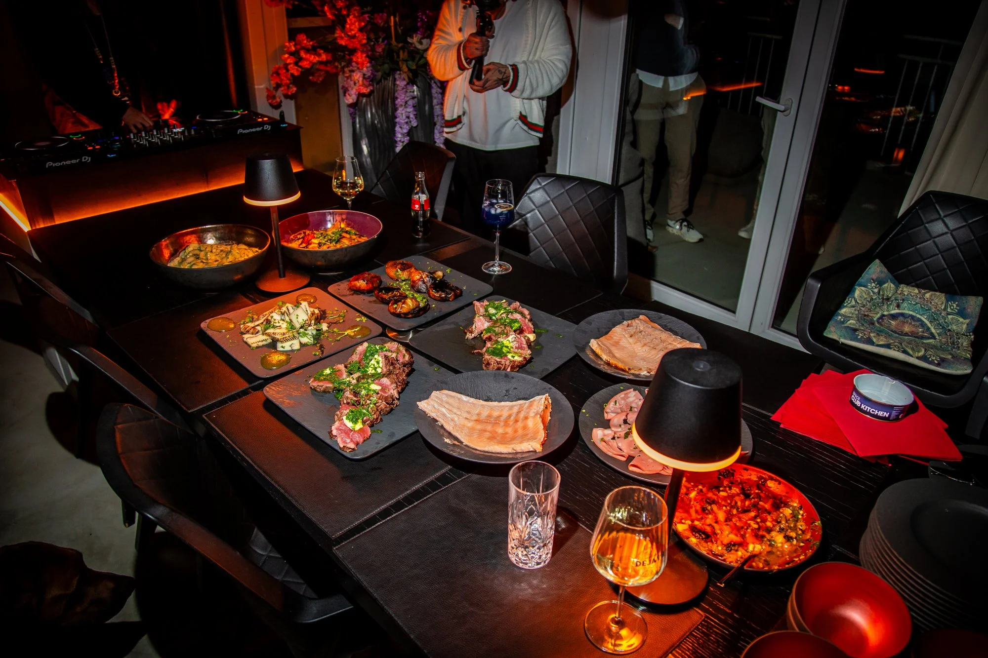 Gedeckte Tafel mit Platten voll Fleisch, Fisch und Salaten im abgedunkelten Raum, dahinter ein DJ-Pult.