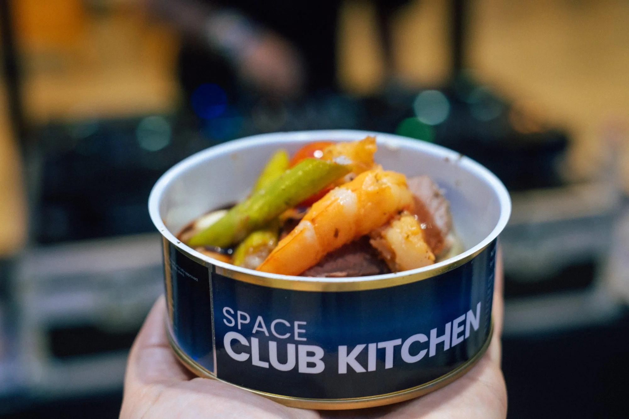 Eine Hand hält eine Dose mit der Aufschrift Space Club Kitchen, gefüllt mit Garnelen und Spargel.