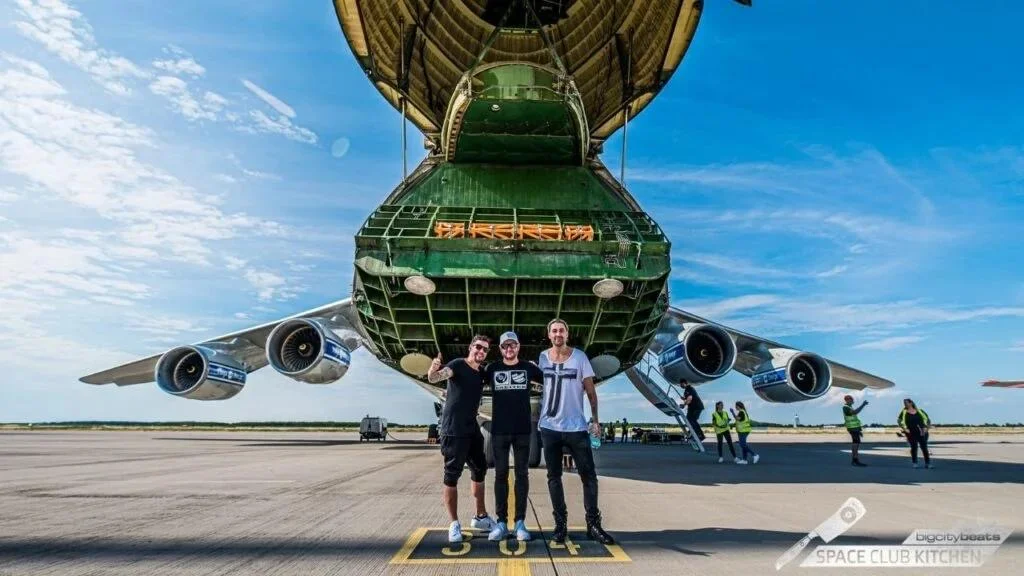Drei Personen auf dem Vorfeld vor einem Frachtflugzeug mit geöffneter Bugklappe
