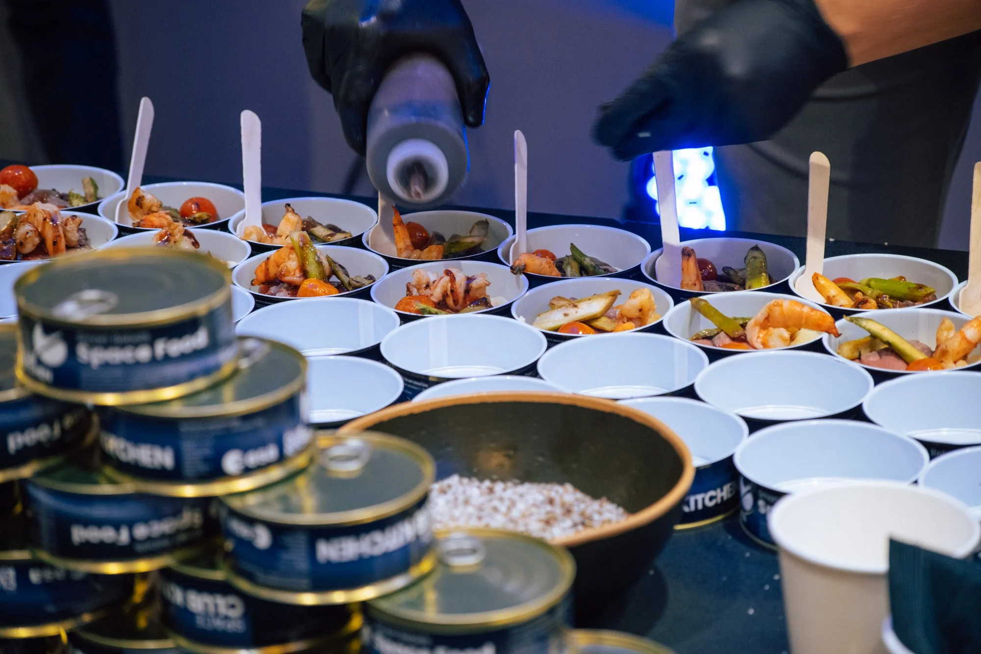 Behandschuhte Hände richten Garnelen und Gemüse in kleinen Bechern neben Space-Food-Dosen an.