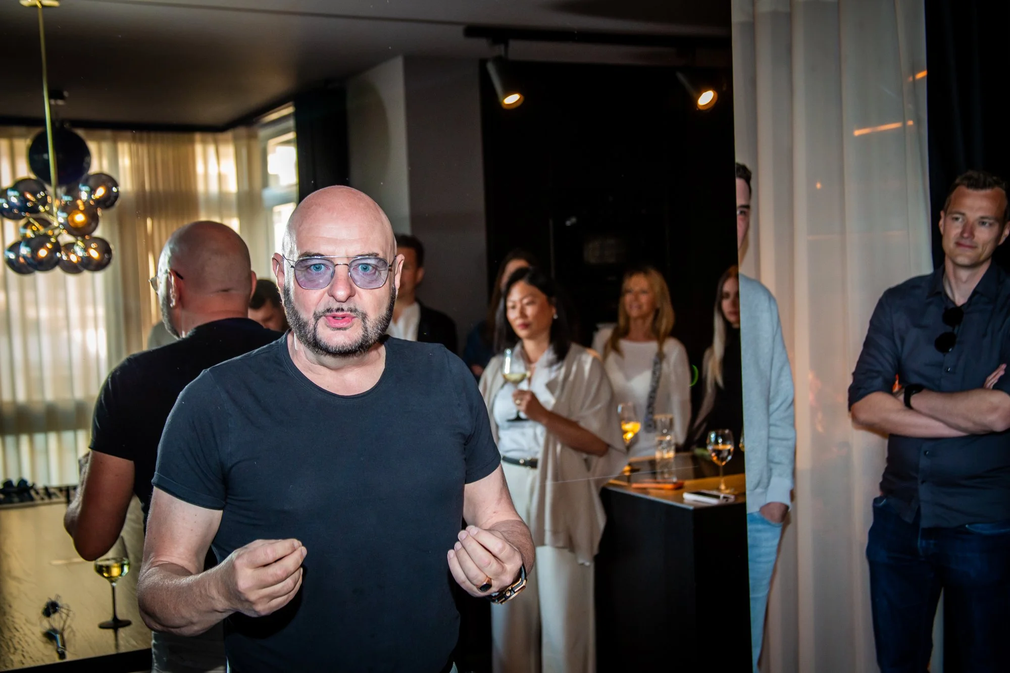 Ein Mann mit Brille und dunklem T-Shirt spricht gestikulierend, dahinter Gäste mit Weingläsern.
