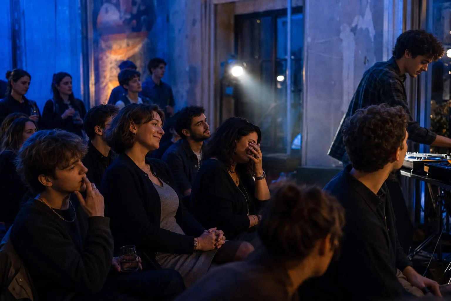 Publikum in blauem Bühnenlicht bei einem Konzert, eine Zuhörerin hält bewegt die Hand vors Gesicht, rechts ein Musiker an einem elektronischen Instrument