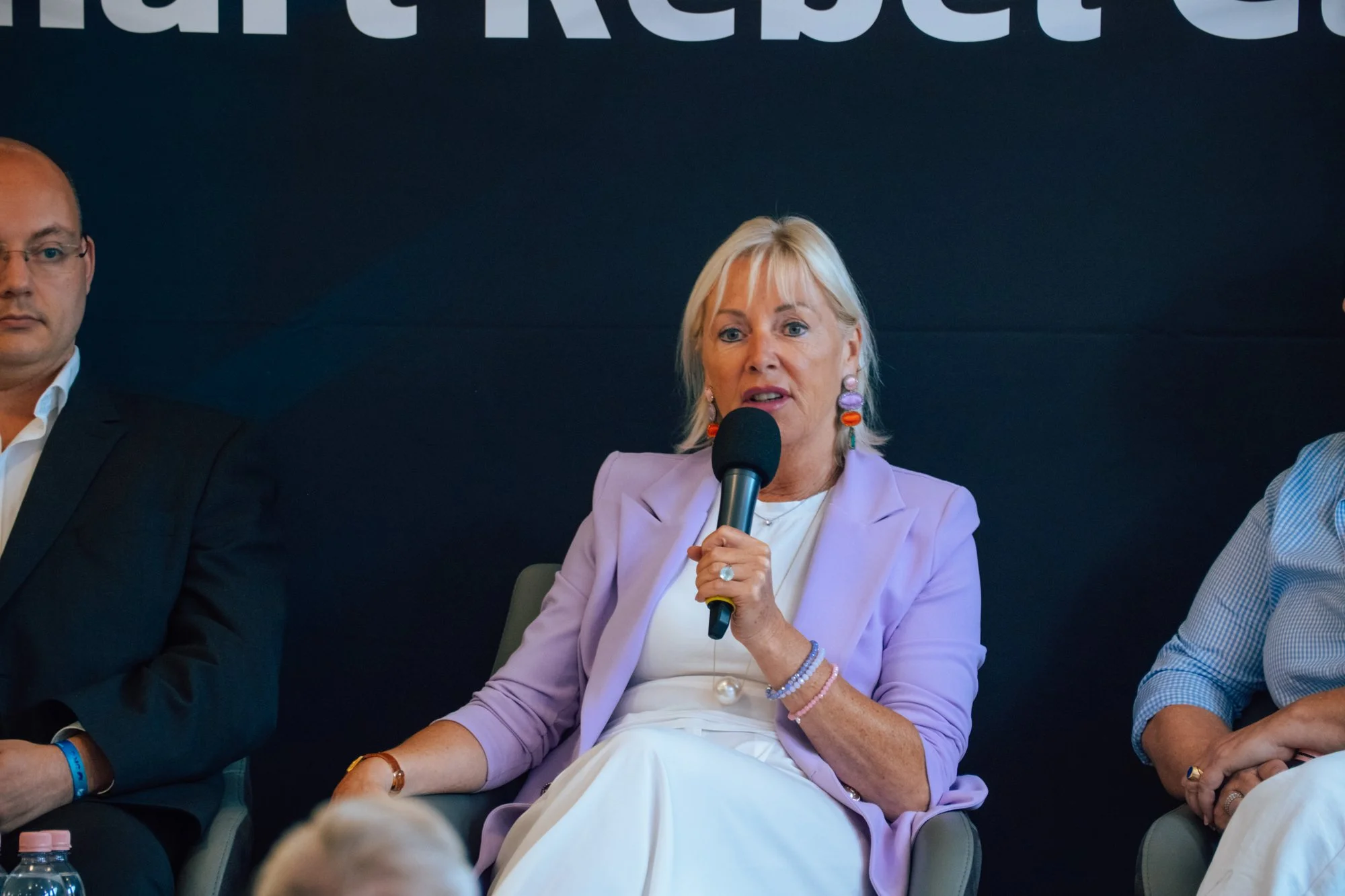 Eine Frau im fliederfarbenen Blazer spricht auf dem Panel ins Mikrofon, neben ihr weitere Gäste.