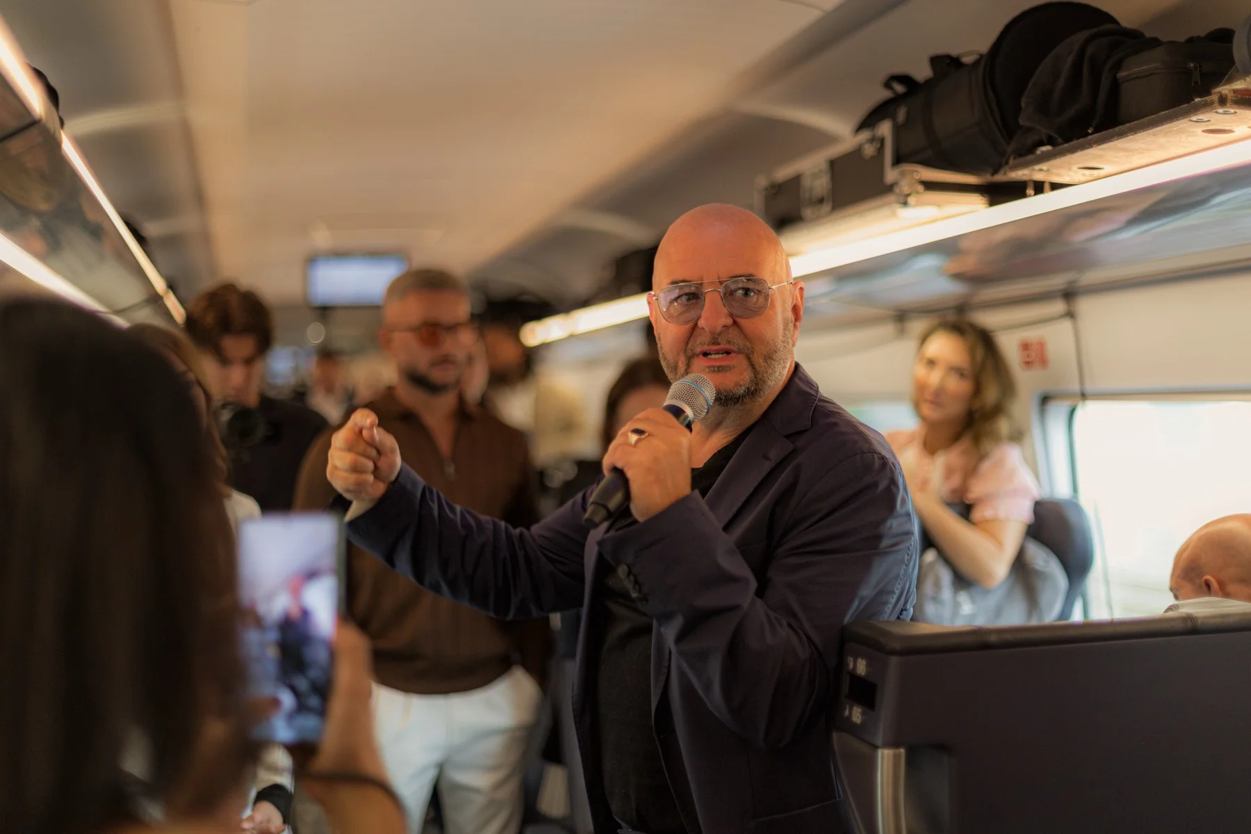 Ein Mann mit Brille spricht gestikulierend ins Mikrofon im Mittelgang des ICE, Mitreisende filmen ihn mit dem Handy.