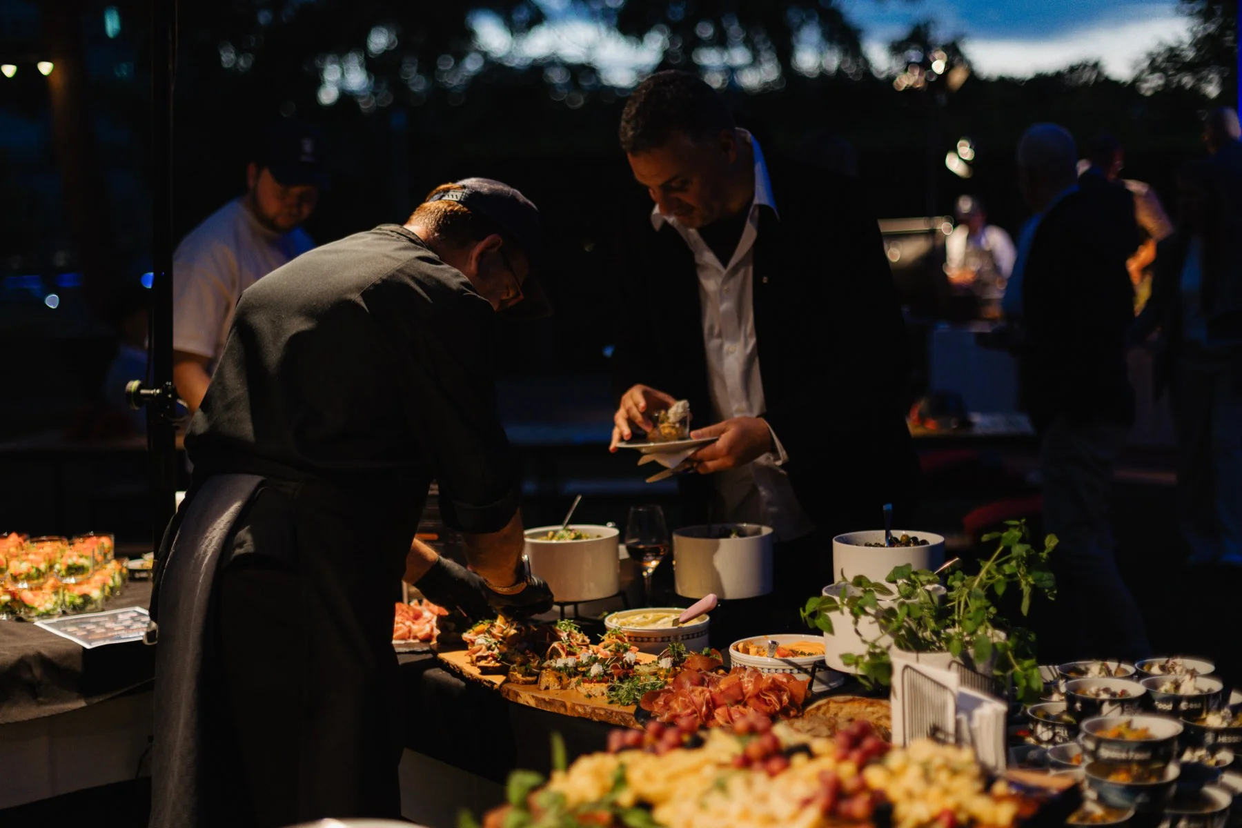 In der Dämmerung richtet ein Koch am Buffet an, ein Gast nimmt sich von einer langen Platte mit Rohschinken, belegten Broten und Kräutern.