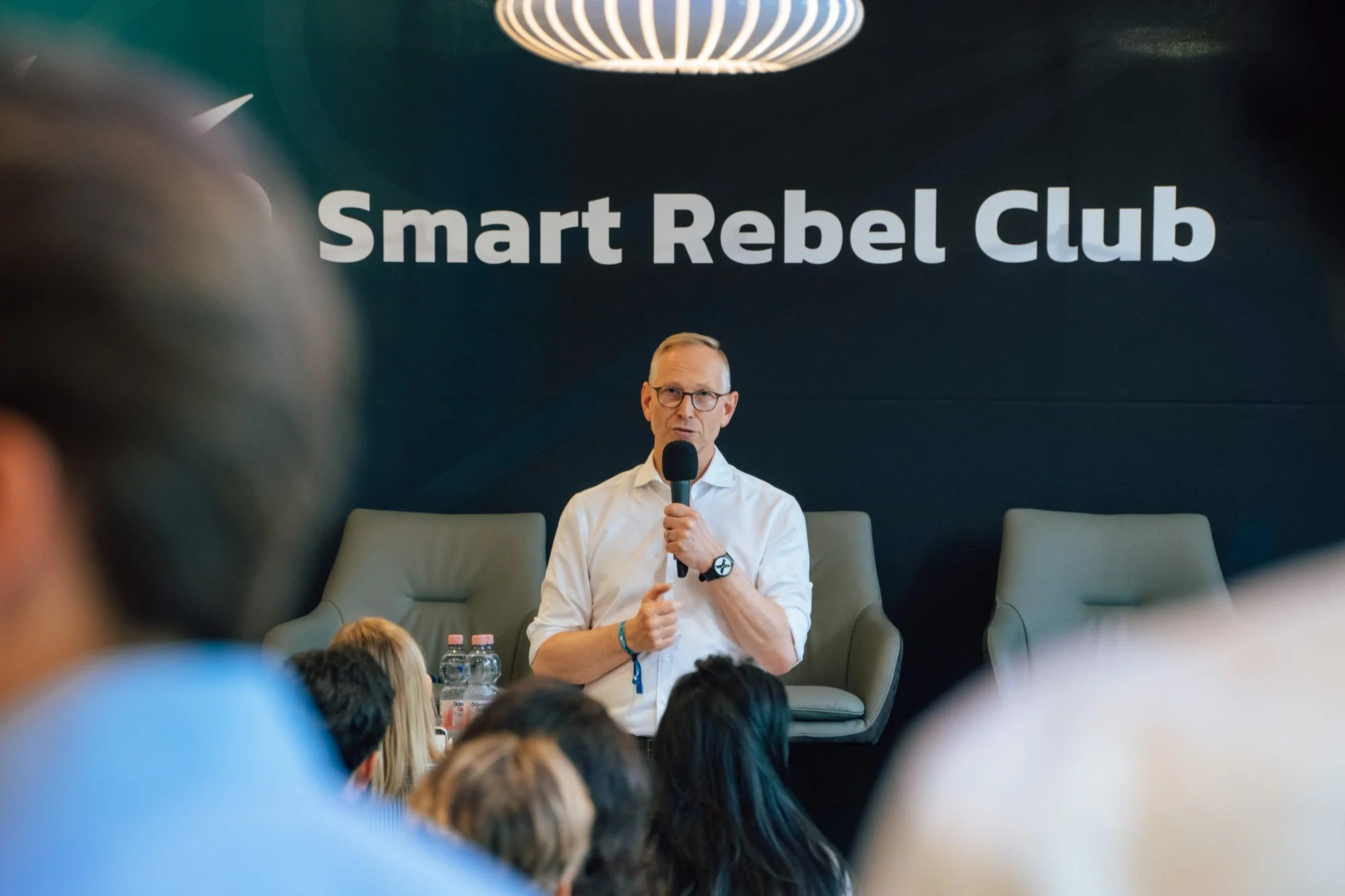Ein Mann im weißen Hemd spricht mit Mikrofon vor der dunklen Smart-Rebel-Club-Wand.