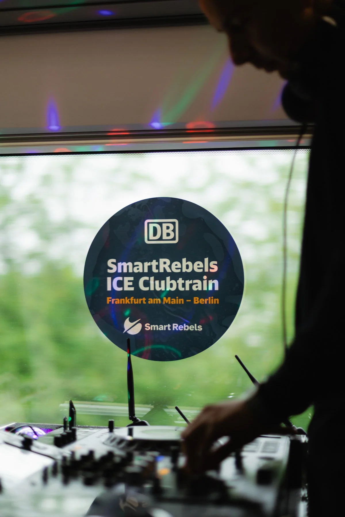 DJ am Mischpult vor dem Zugfenster, darauf ein runder Aufkleber mit dem DB-Logo und der Aufschrift SmartRebels ICE Clubtrain Frankfurt am Main – Berlin.