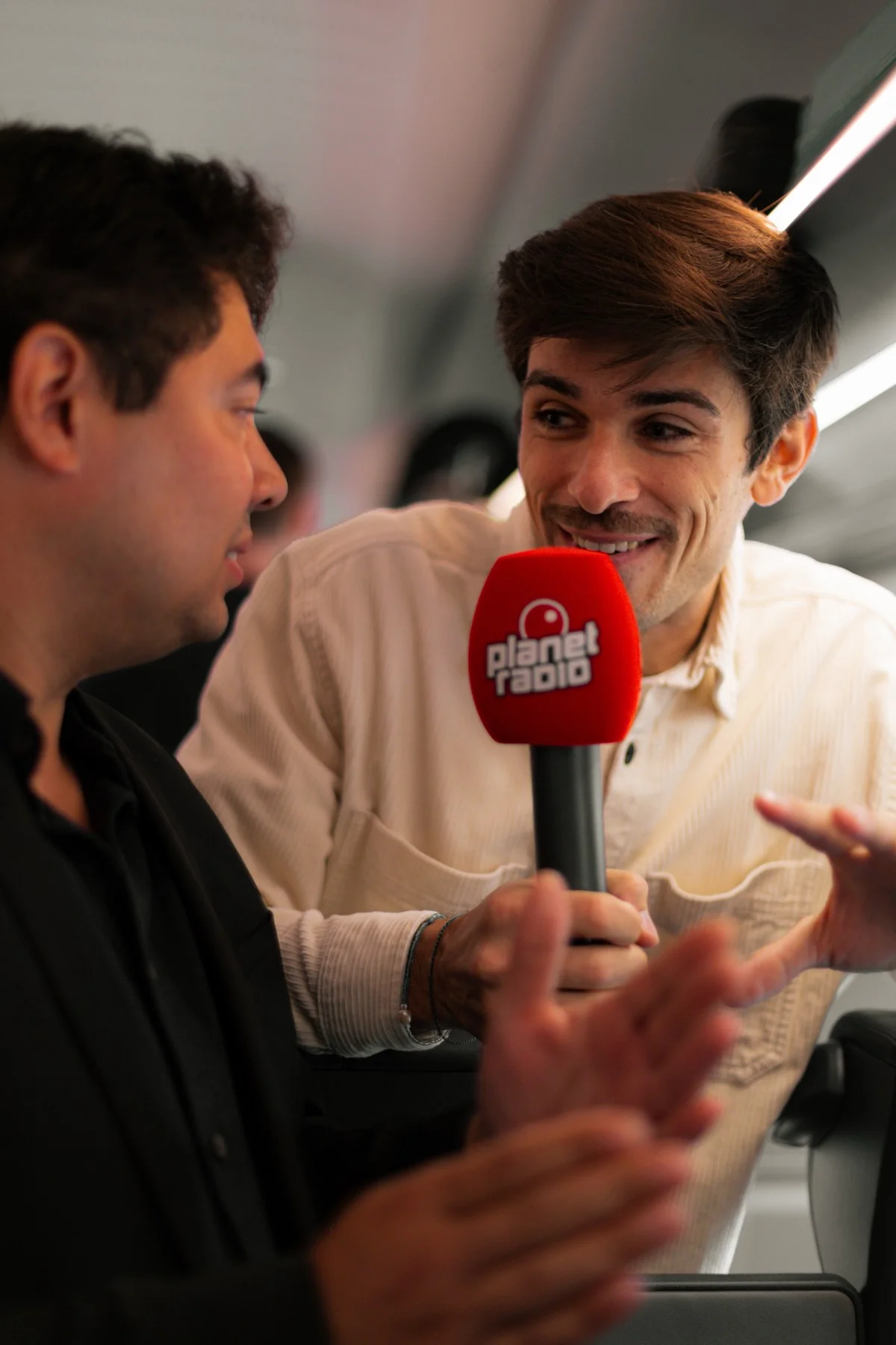 Zwei Männer im Gespräch im Zug, einer hält ein rotes Mikrofon mit dem Logo von Planet Radio.