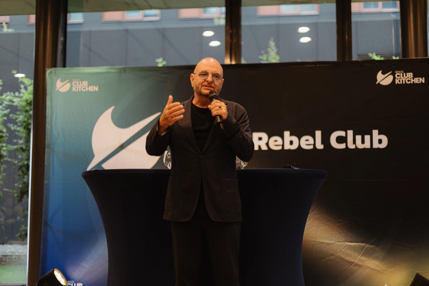 Ein Mann spricht mit Mikrofon vor einer dunklen Wand mit dem Schriftzug Smart Rebel Club und dem Logo der Space Club Kitchen.