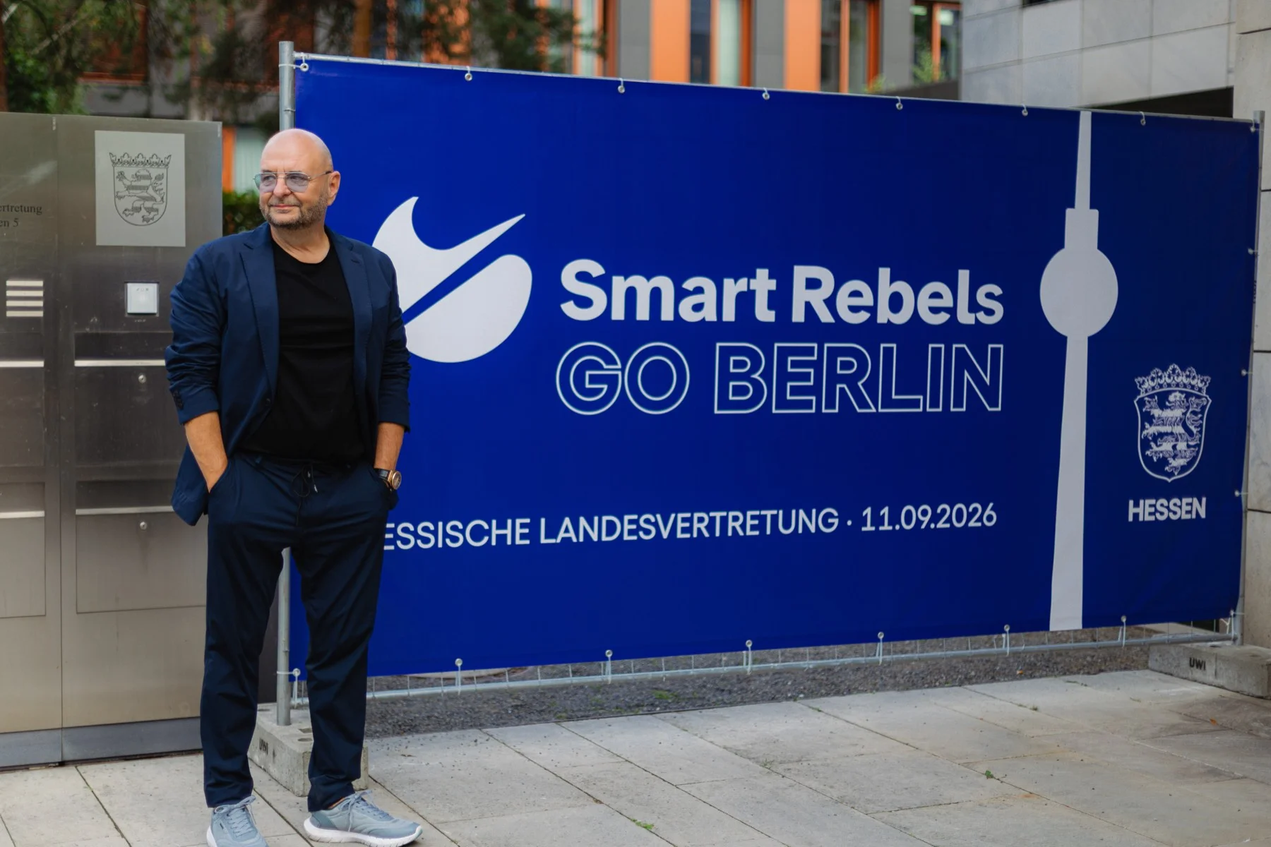 Ein Mann steht vor einem blauen Banner mit der Aufschrift Smart Rebels Go Berlin, Hessische Landesvertretung, 11.09.2026 und dem hessischen Wappen.