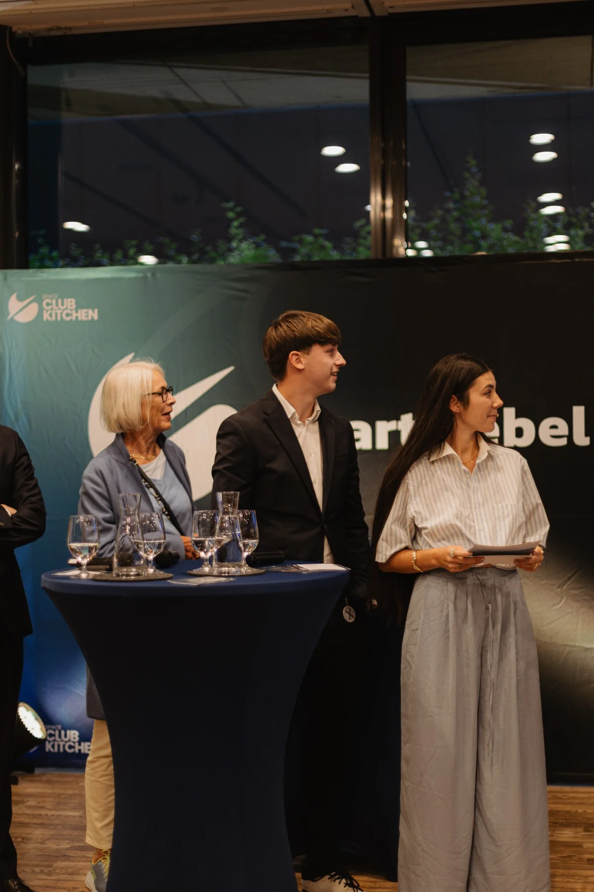Eine ältere Frau, ein junger Mann im dunklen Anzug und eine junge Frau mit Karten stehen an Stehtischen vor der Wand des Smart Rebel Clubs.