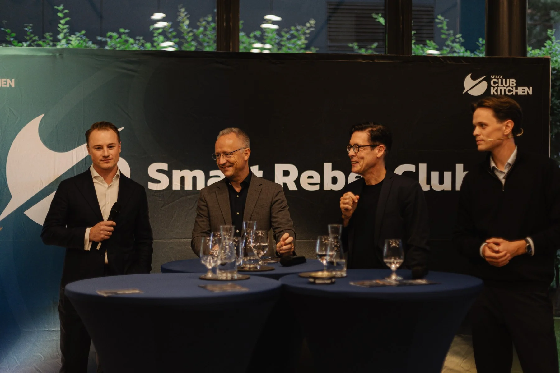 Vier Männer stehen an Stehtischen mit Wassergläsern, einer hält ein Mikrofon, vor einer dunklen Wand mit der Aufschrift Smart Rebel Club und dem Logo der Space Club Kitchen.