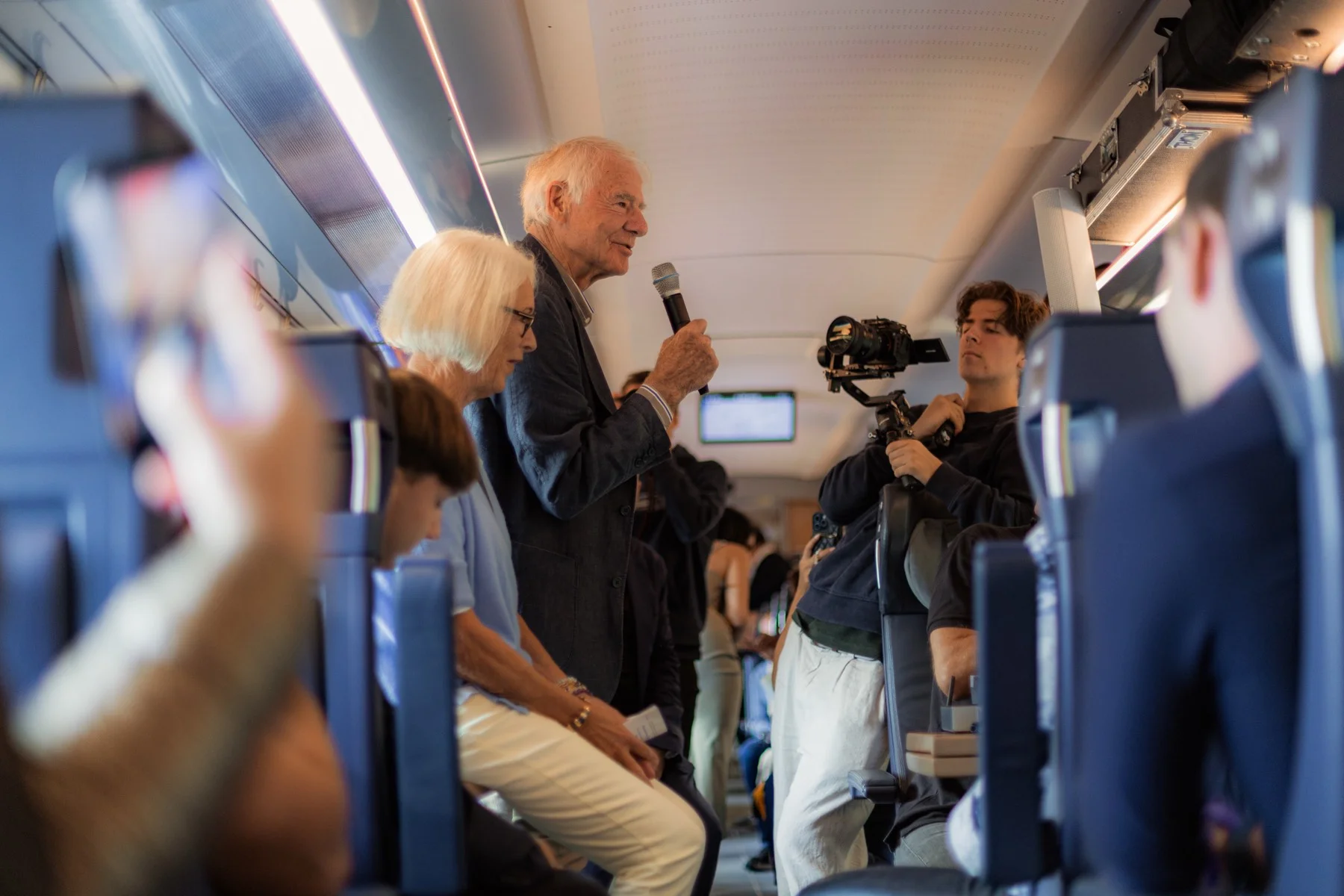 Ein älterer Herr mit weißem Haar spricht stehend ins Mikrofon im Zugabteil, rechts filmt ihn ein Kameramann mit einer Kamera auf einem Gimbal an einem Griffstativ.
